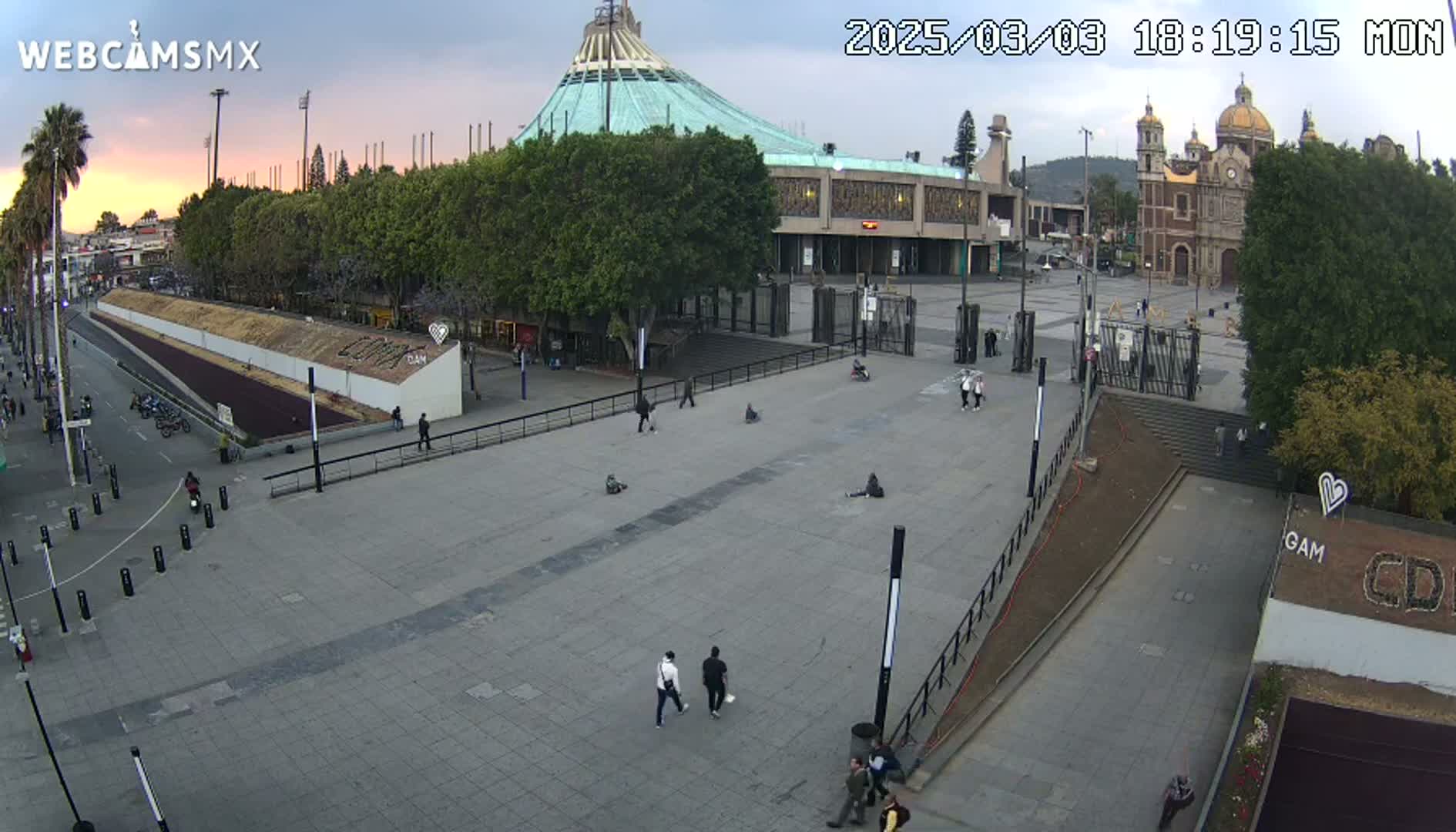 A large plaza with a few pedestrians, situated in front of a large basilica under a partly cloudy sunset sky.