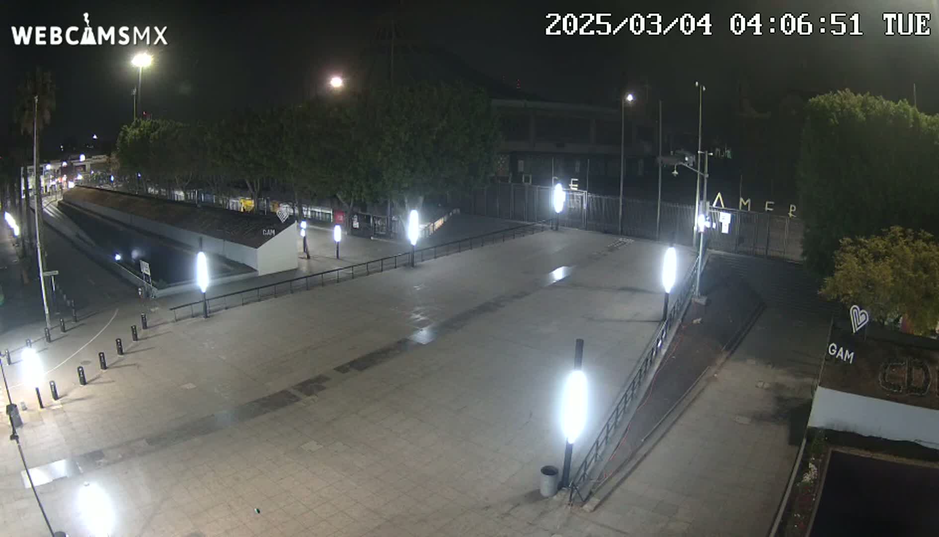 A nighttime, outdoor view of a paved plaza with several lampposts, some trees, and a gated area in the background.