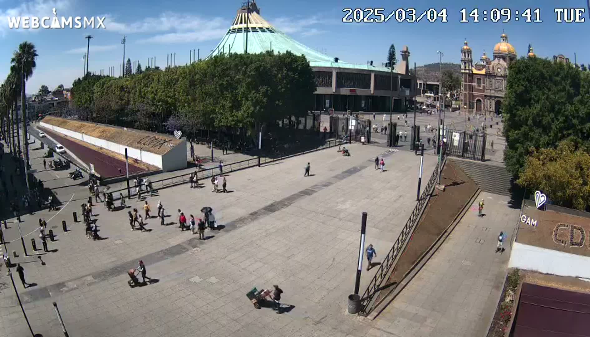 A wide outdoor shot on a sunny day shows a large plaza with a few people walking around, in front of a large basilica and a modern building.