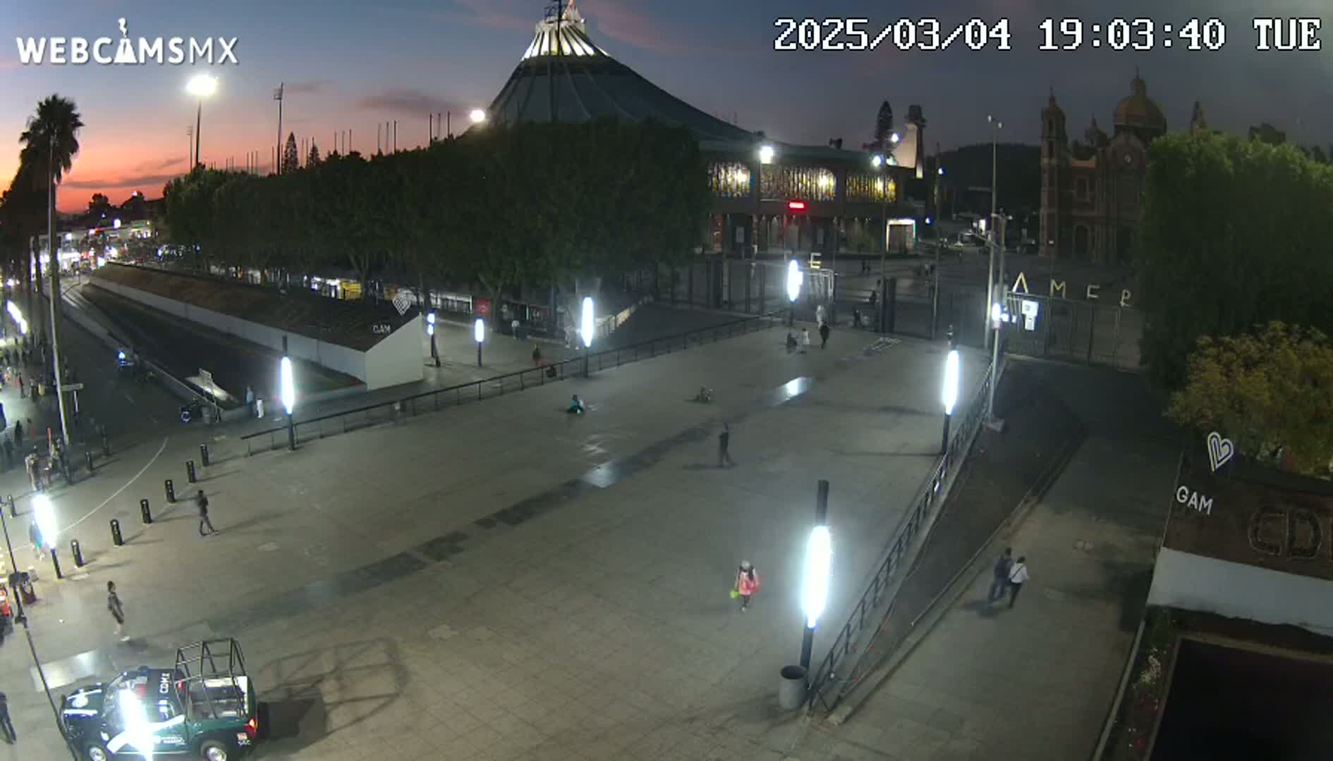 A nighttime, dimly lit plaza with a large basilica in the background, and a few people walking around.