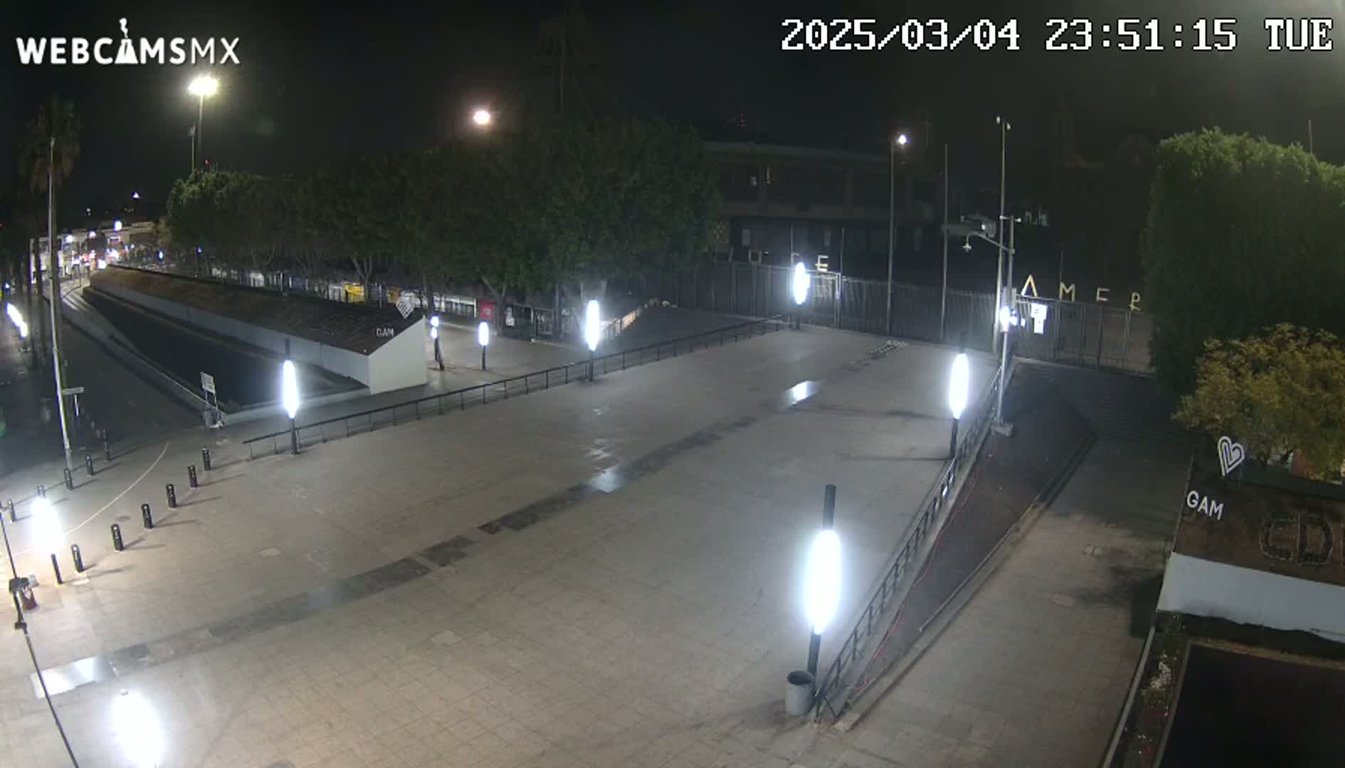 A nighttime view of an empty plaza with several lights illuminating a paved area, surrounded by trees and buildings under a clear sky.