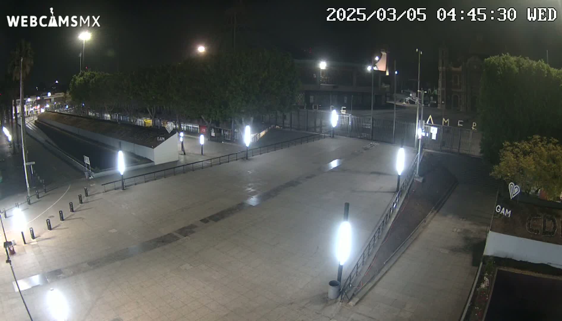 A nighttime, outdoor view of a paved plaza with several lampposts, some trees, and a gated entrance to a building.