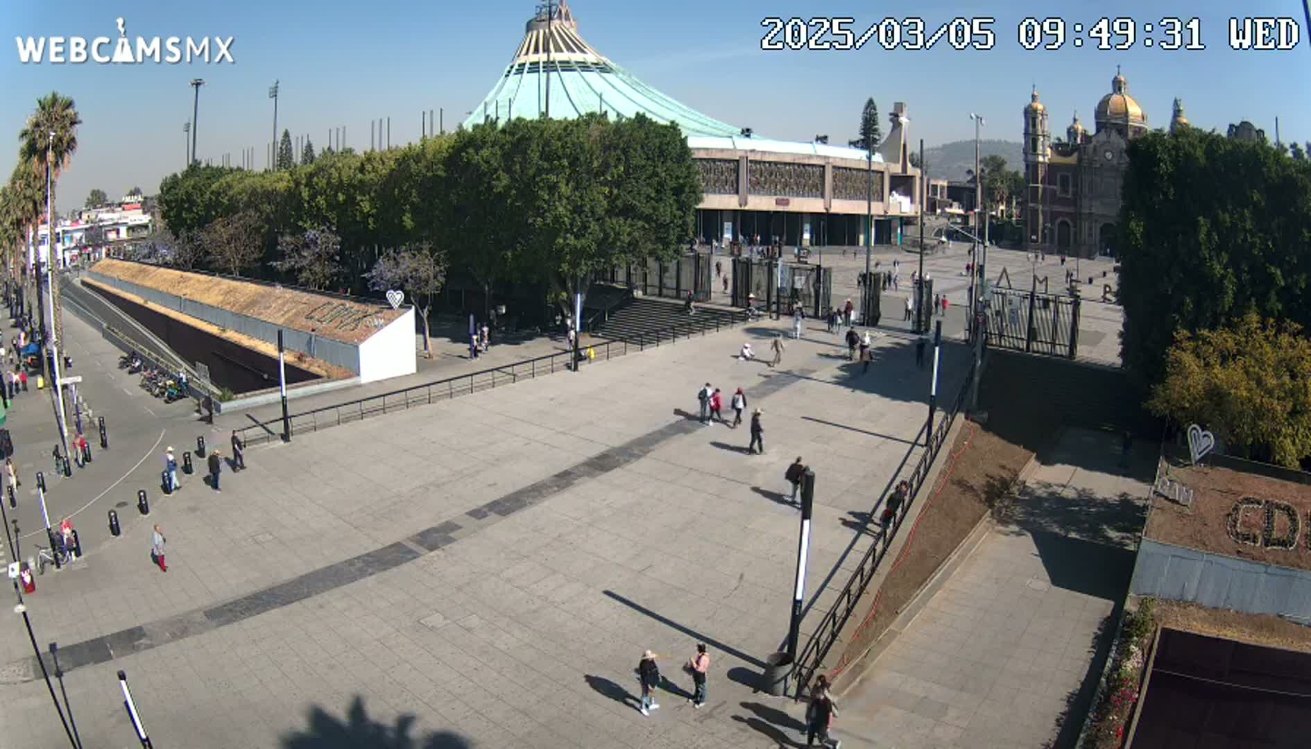 A large plaza with a few people walking, surrounded by buildings including a large domed structure and a church, on a sunny day.