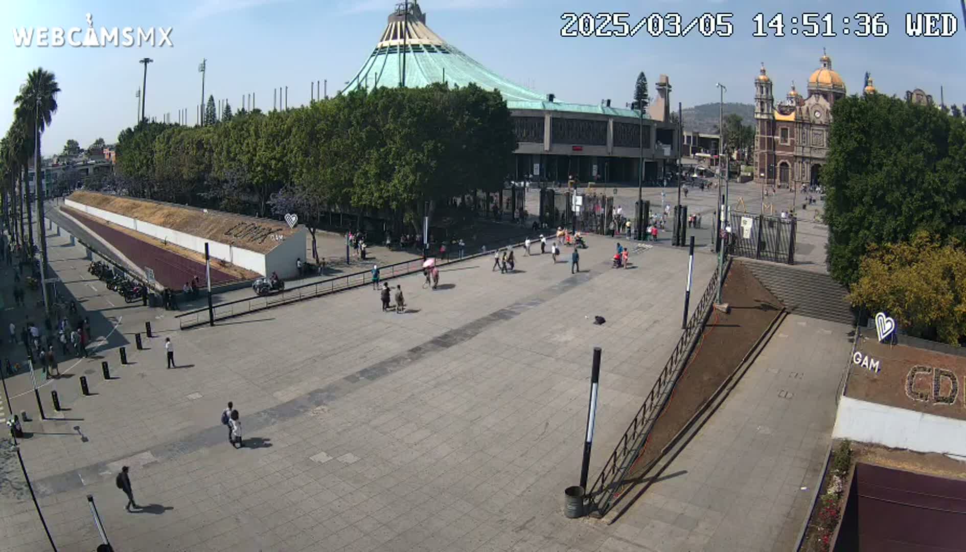 A wide, paved plaza with a few people walking around, in front of a large basilica and a modern building under a sunny sky.