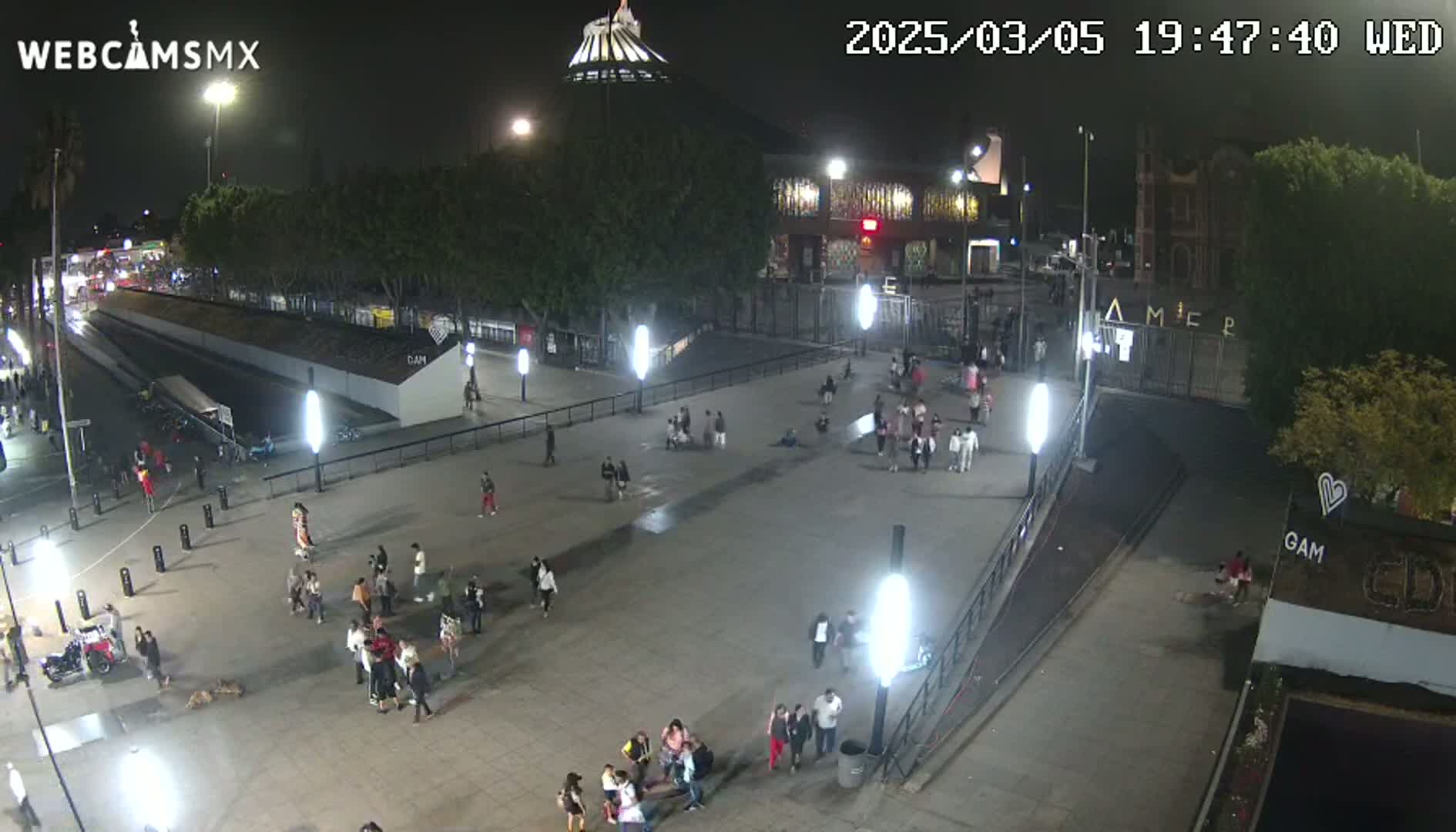 A nighttime, outdoor view of a plaza with numerous people gathered, illuminated by artificial lights, with buildings and trees surrounding the area.