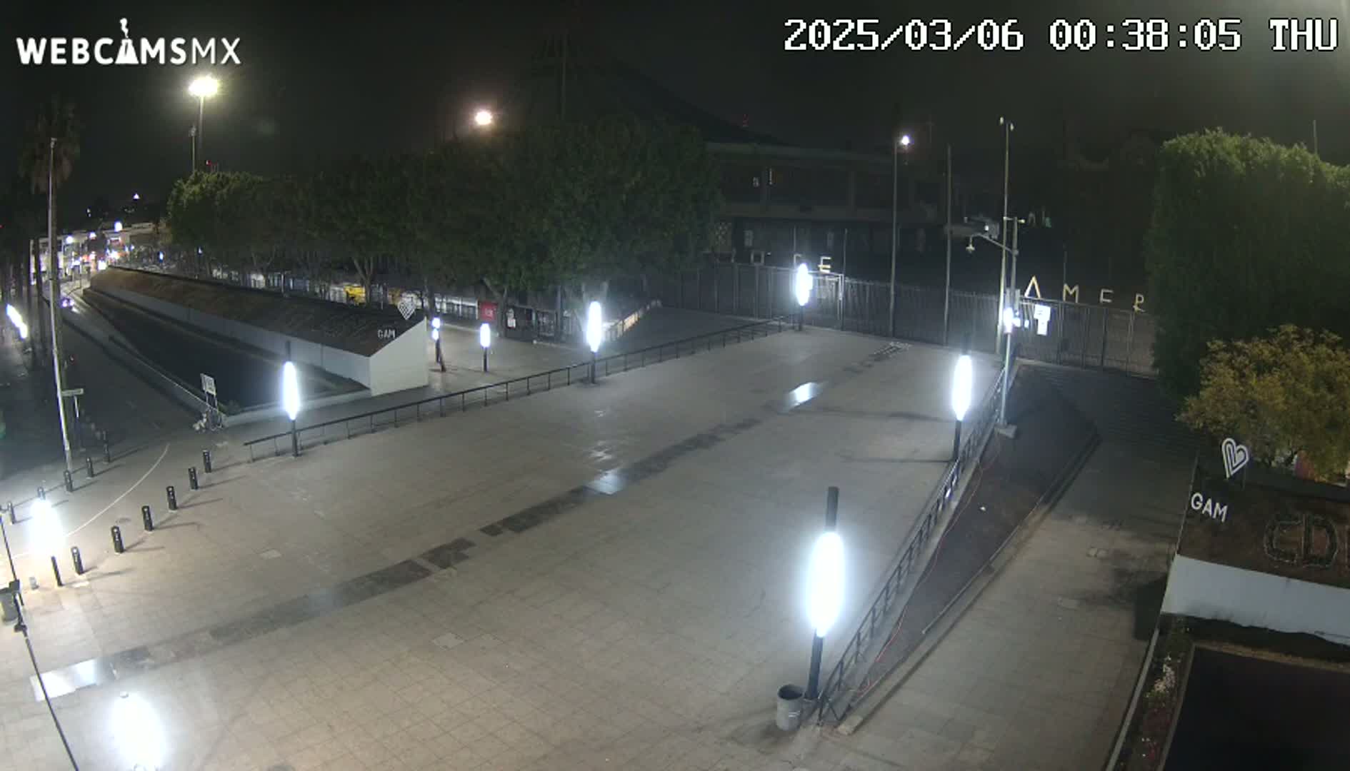 A nighttime view of an empty plaza with several light posts, trees, and a gated area beyond.