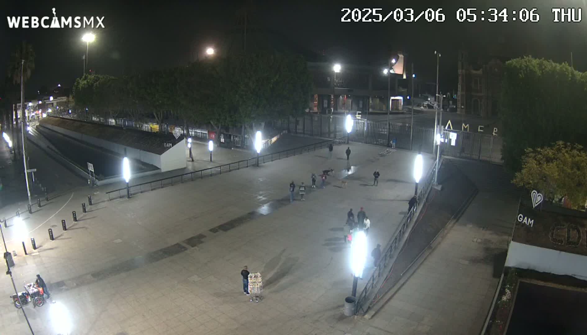 A nighttime, outdoor view of a mostly empty plaza with several people standing in small groups, illuminated by bright lights, near a gated area with trees and buildings in the background.