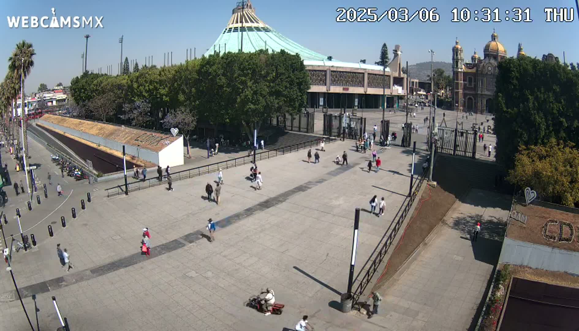 A plaza with a large basilica in the background, under sunny skies, has several people walking around.