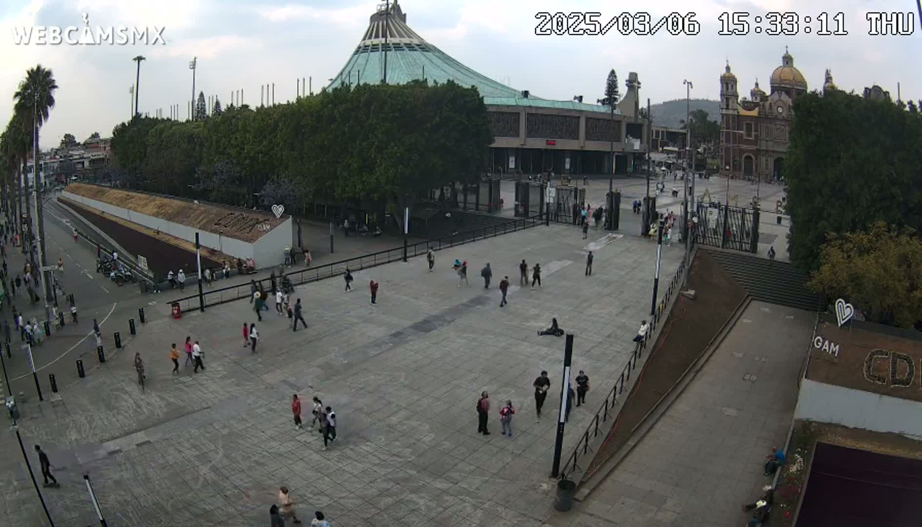 A large plaza with a basilica and a modern building in the background, under a partly cloudy sky, is populated by a scattering of people.