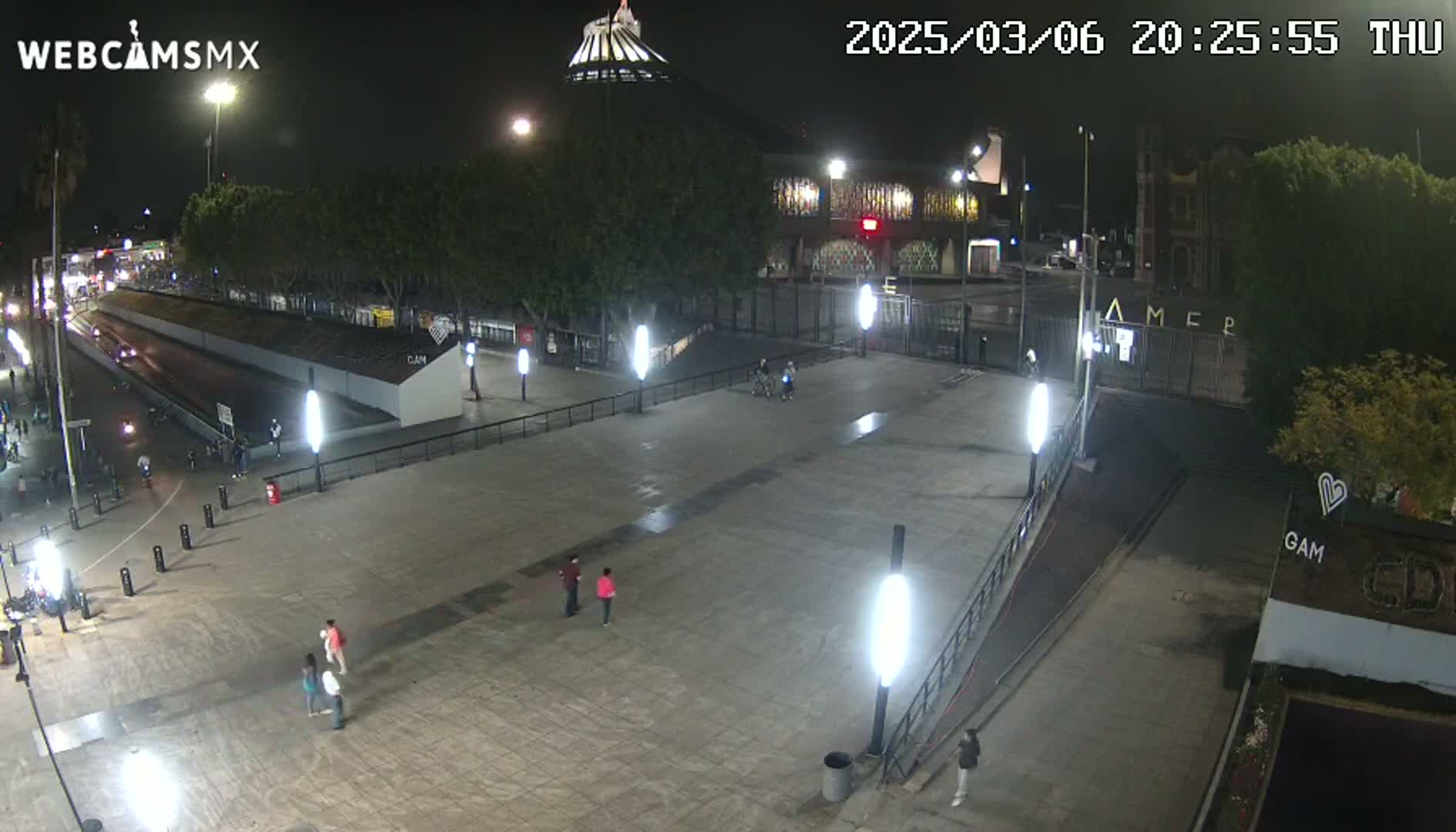 A nighttime, mostly empty plaza with several people walking around, lit by bright lights, and featuring buildings and trees in the background.