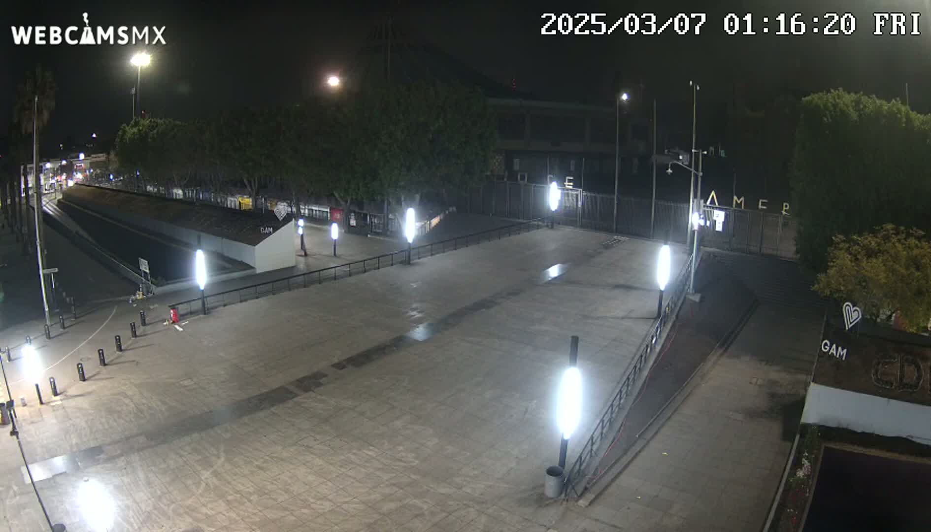 A nighttime, paved plaza with several lampposts and trees surrounding a gated area.