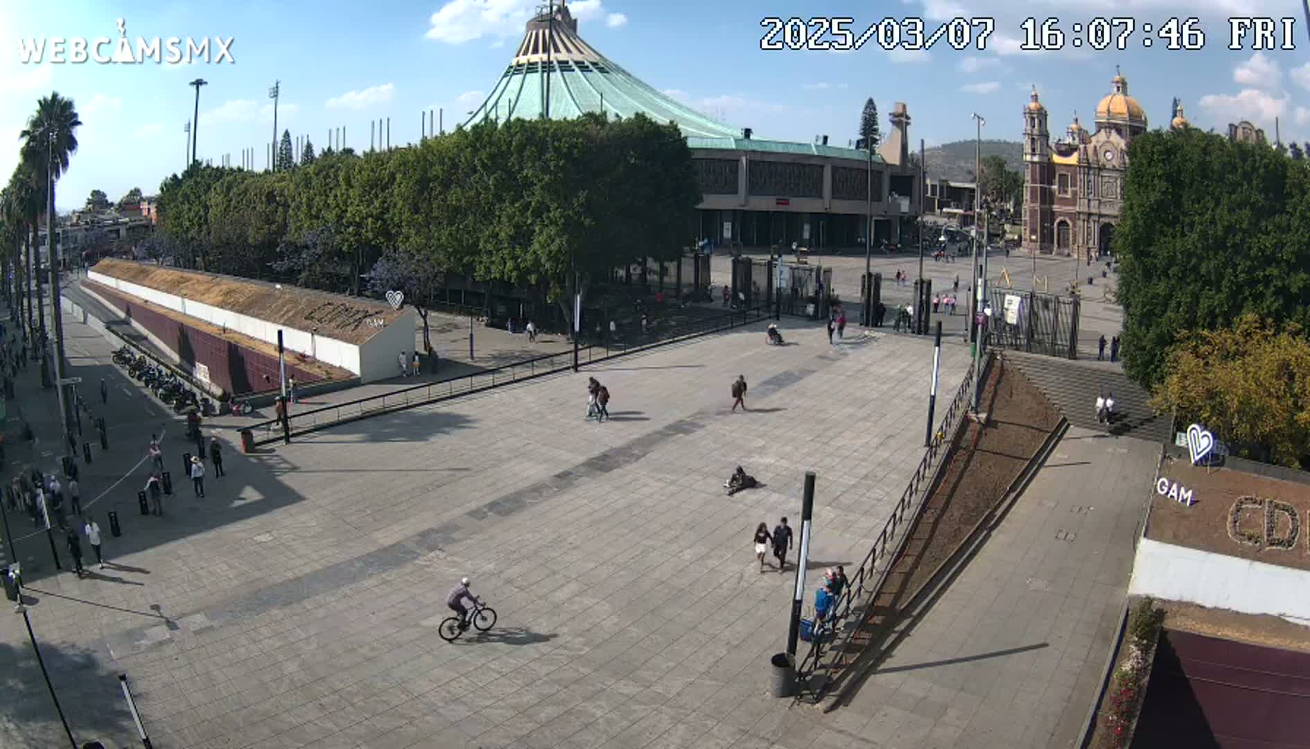 A large plaza with a few people walking and biking, in front of a large basilica and a smaller, modern building under a sunny sky.