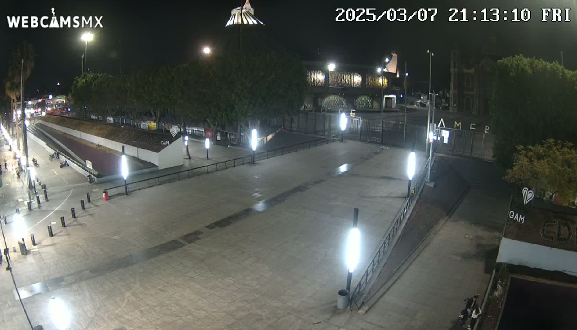 A nighttime, paved plaza with several lights and sparse pedestrian traffic, near a gated area and some buildings.