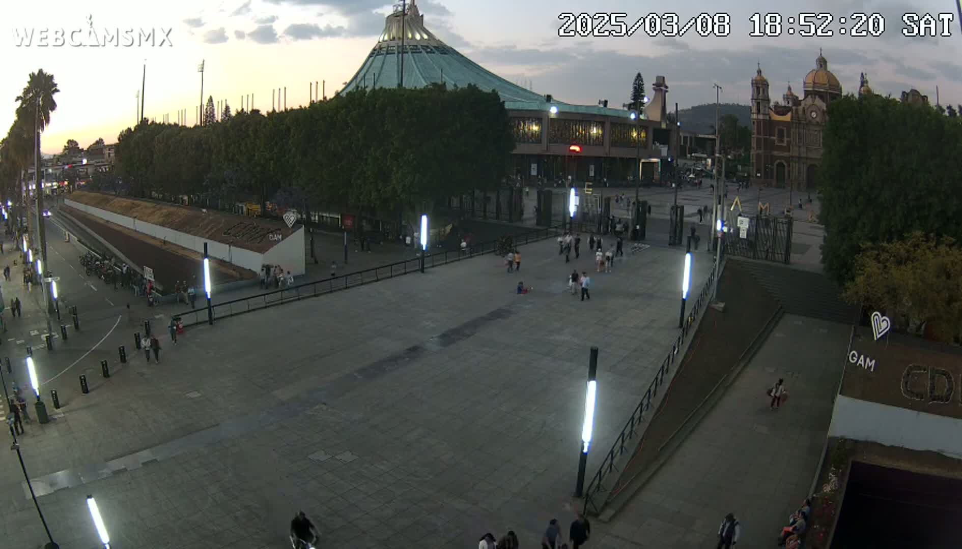 A wide, paved plaza at dusk features a large basilica and a modern building, with several people scattered throughout the area under a mostly clear sky.