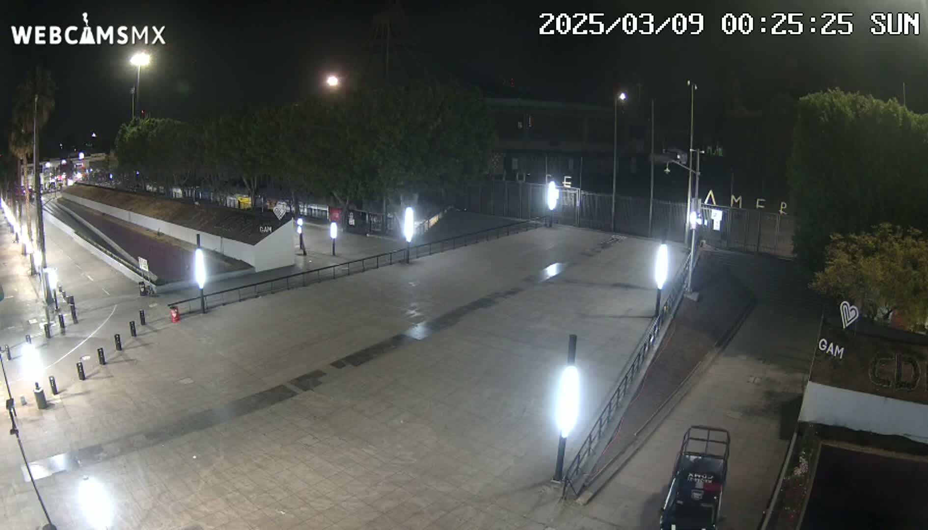 A nighttime view of an empty plaza with several tall light posts illuminating the paved area, surrounded by trees and buildings.