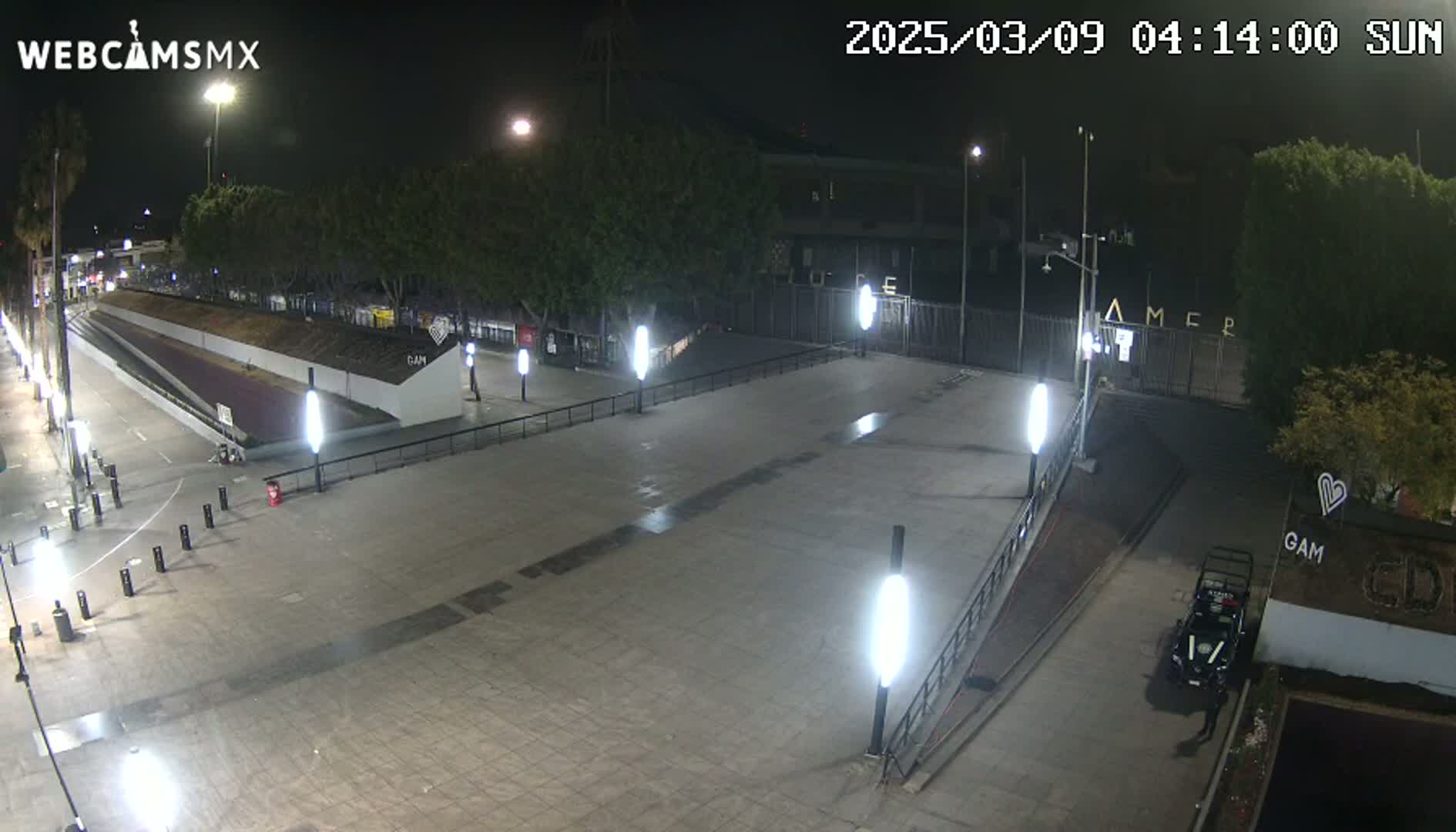 A nighttime view of an empty plaza with several lights illuminating a paved area leading to a gated entrance, under clear skies.