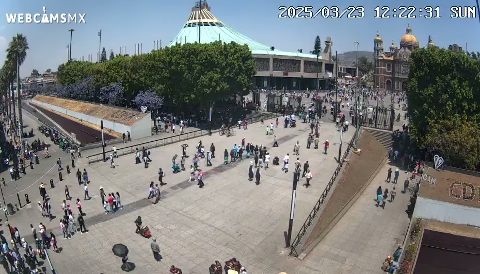 A large plaza with many people, a basilica, and surrounding trees under sunny skies.