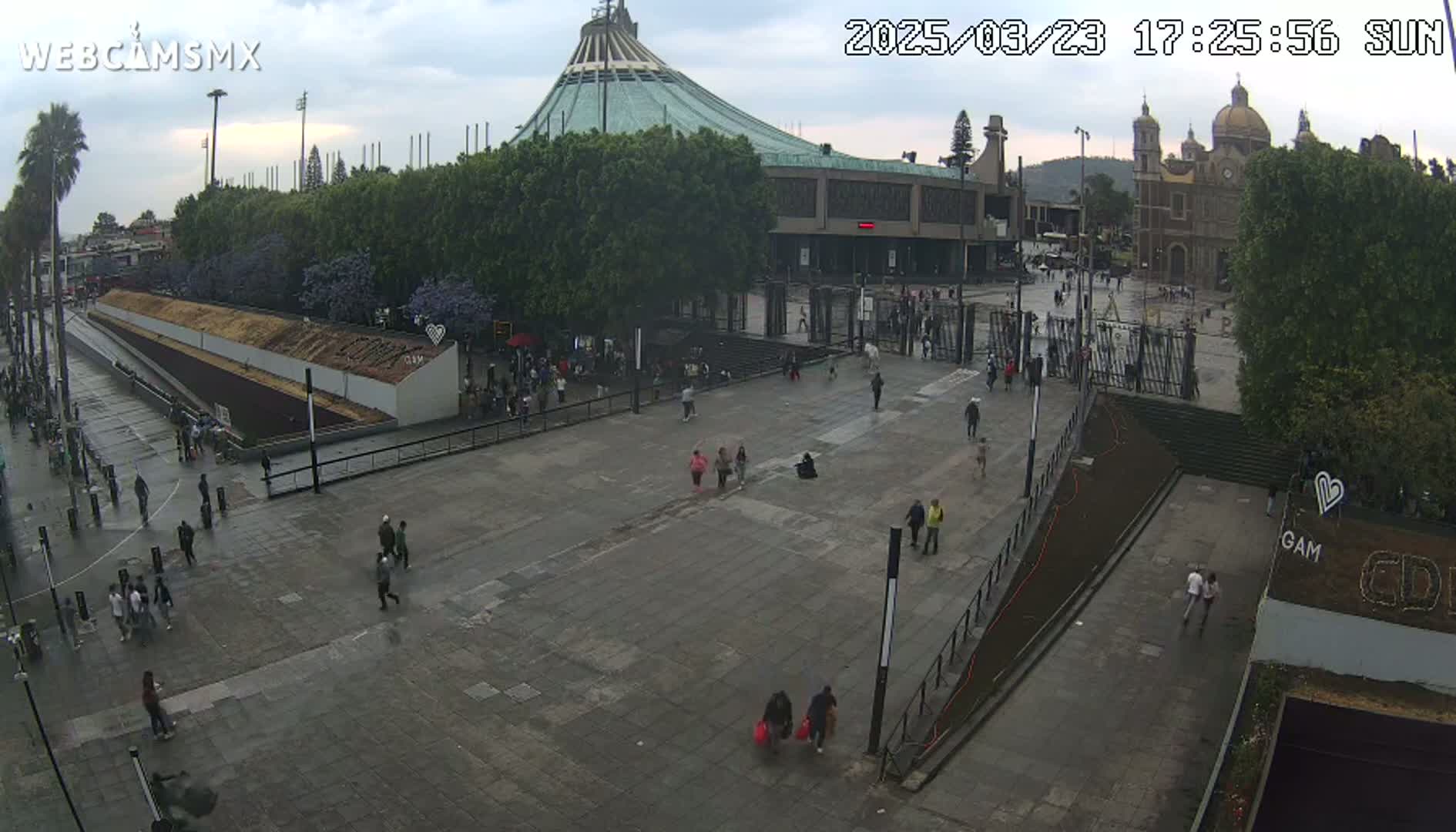 A large plaza with several people walking around, situated in front of a large basilica and a modern building under an overcast sky.