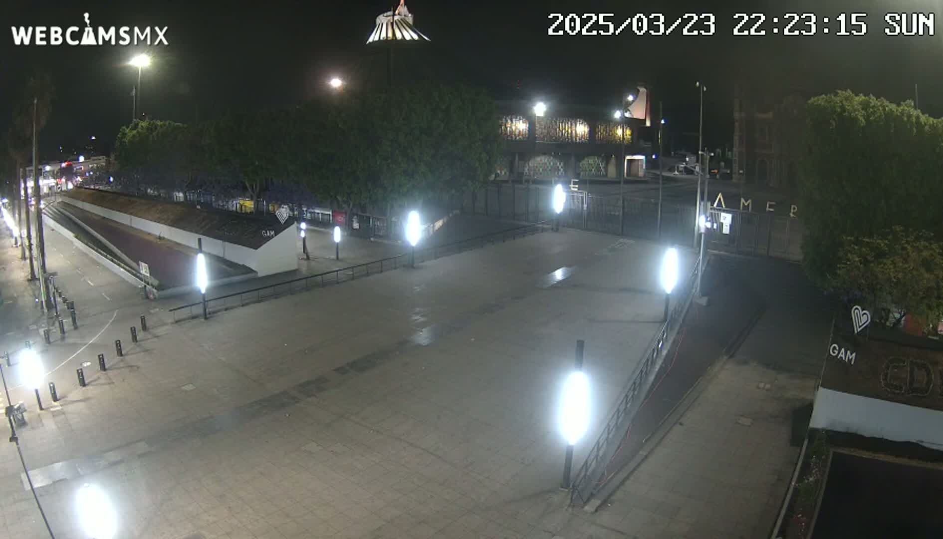 A nighttime, outdoor view of a mostly empty plaza with several lights and some trees, illuminated by artificial light.