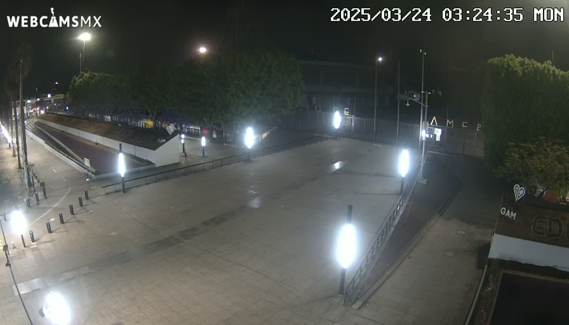 A nighttime, paved plaza with several lampposts is shown, with a sloped walkway leading to a gated area beyond some trees.