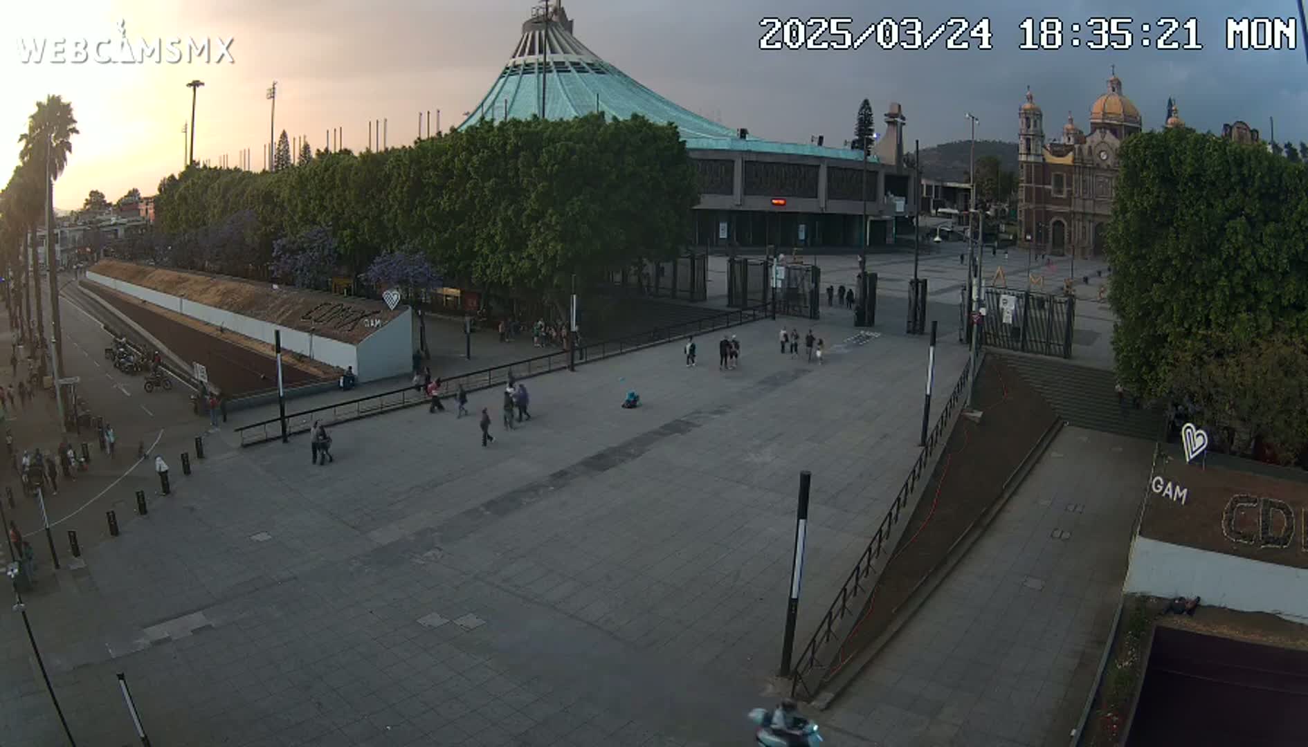 A wide outdoor shot at dusk shows a large plaza with a few people walking around, in front of a large building and a church.