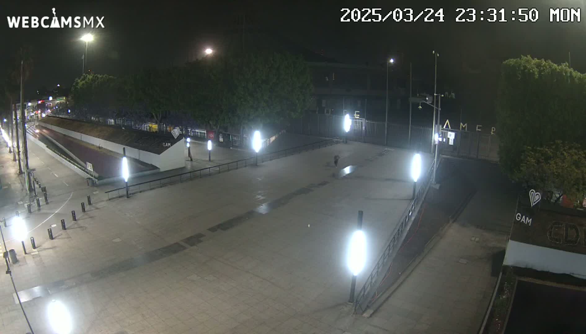 A nighttime view of a paved plaza with several lights, trees, and a ramp leading to a gated area.