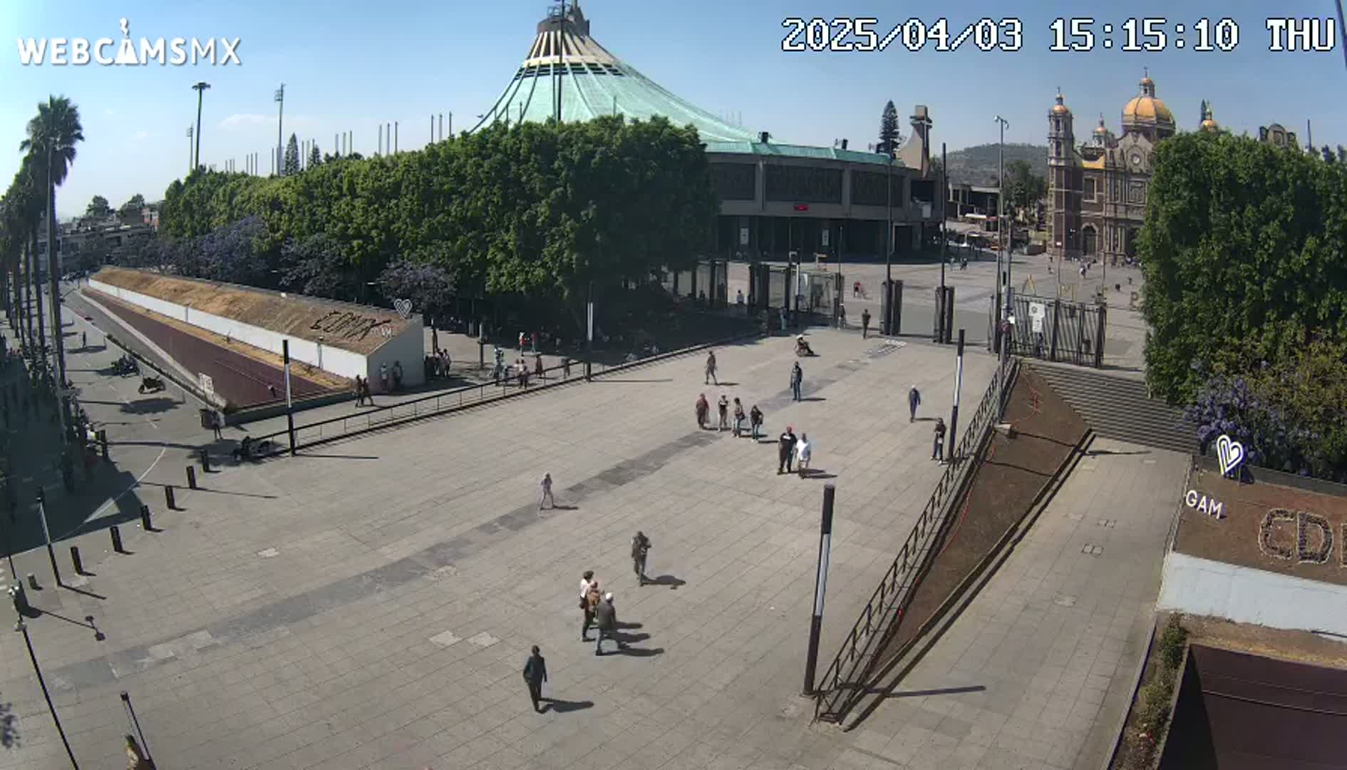 A large plaza with a few people walking, situated in front of a large basilica and a modern building under a clear, sunny sky.