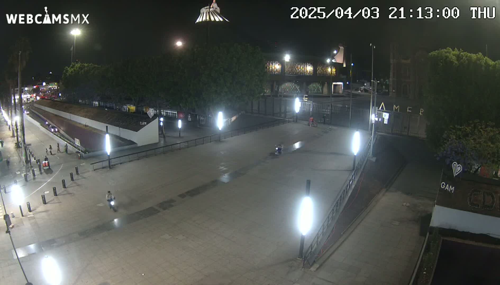 A nighttime, outdoor view of a plaza with several people walking and riding scooters, illuminated by streetlights under a clear sky.