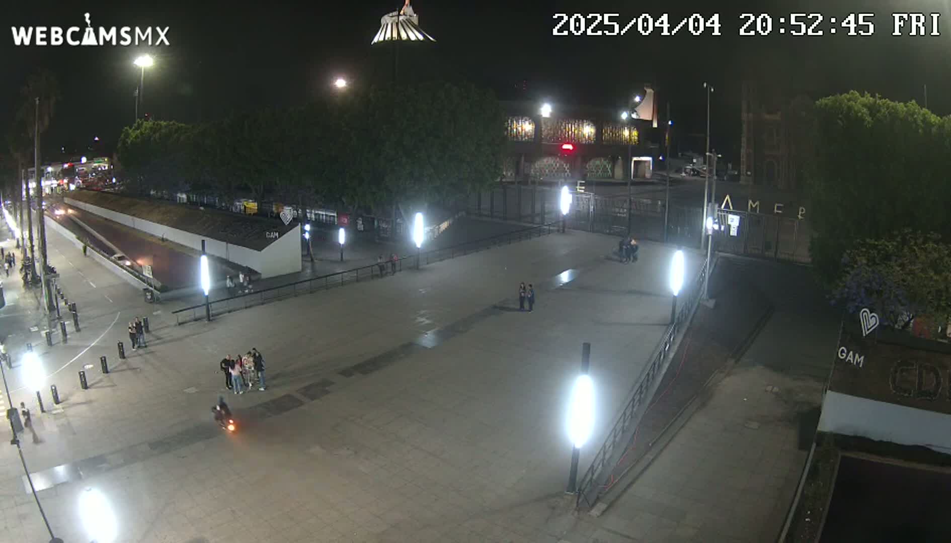 A nighttime, outdoor view of a plaza with several people, illuminated by bright lights, near a building with graffiti.