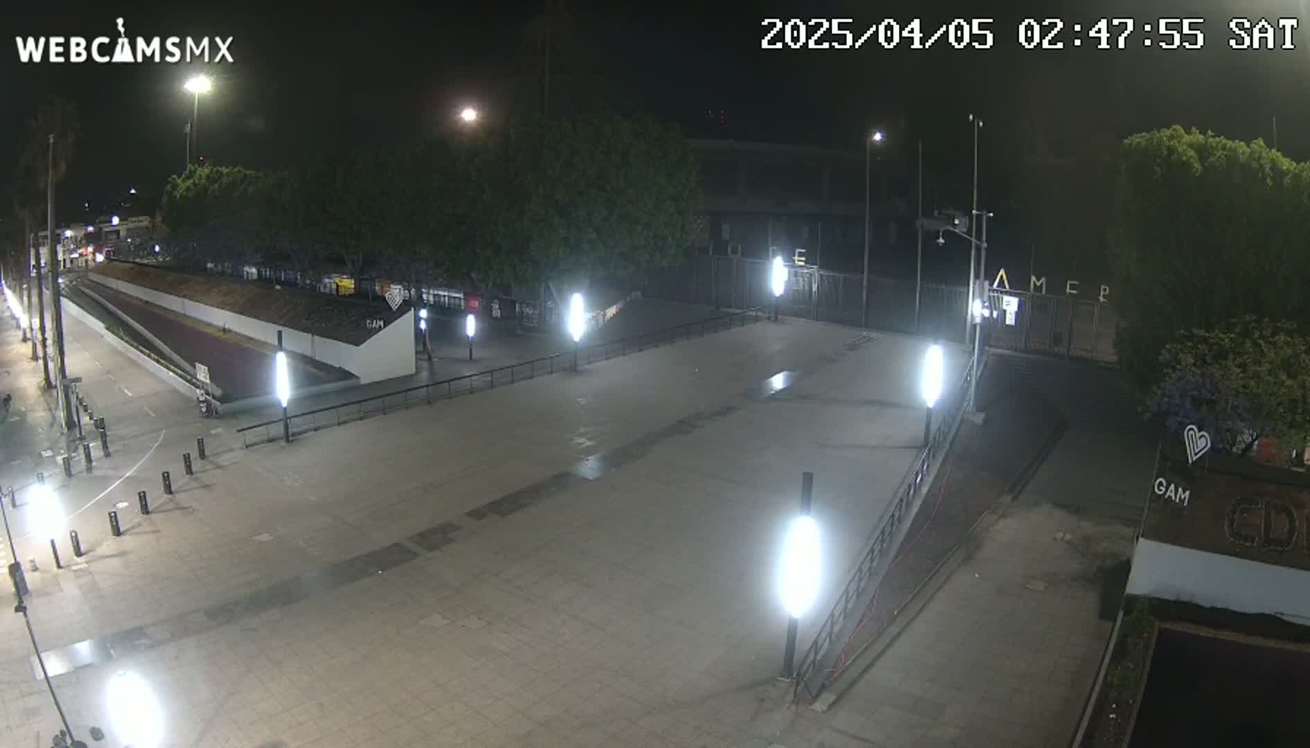 A nighttime, brightly lit plaza with a paved pathway and several trees is seen, with some buildings and fencing in the background.