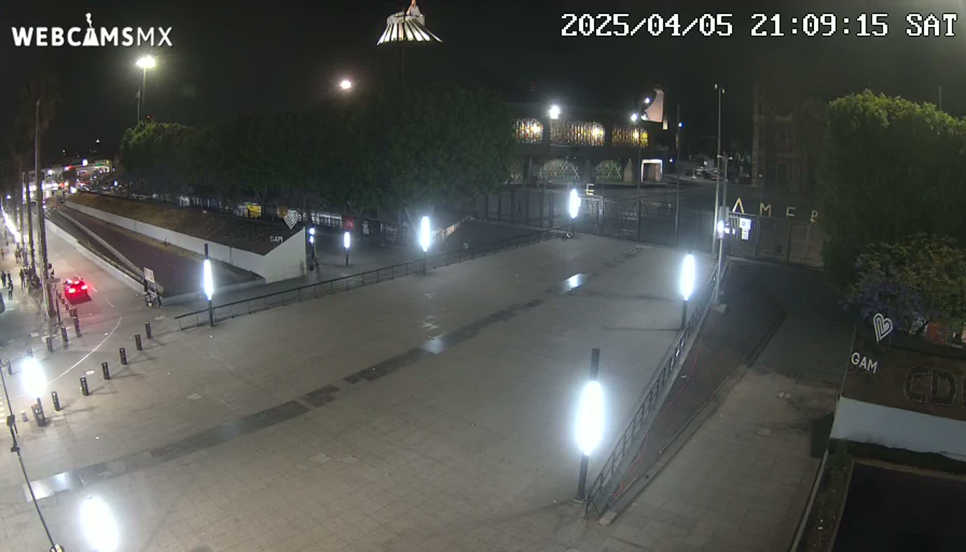 A nighttime, outdoor view of an empty plaza with several bright lights, a road with one car, and buildings in the background.