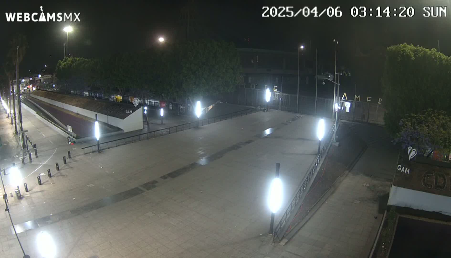 A nighttime view of an empty plaza with several illuminated lampposts, a walkway, and a gated area beyond.