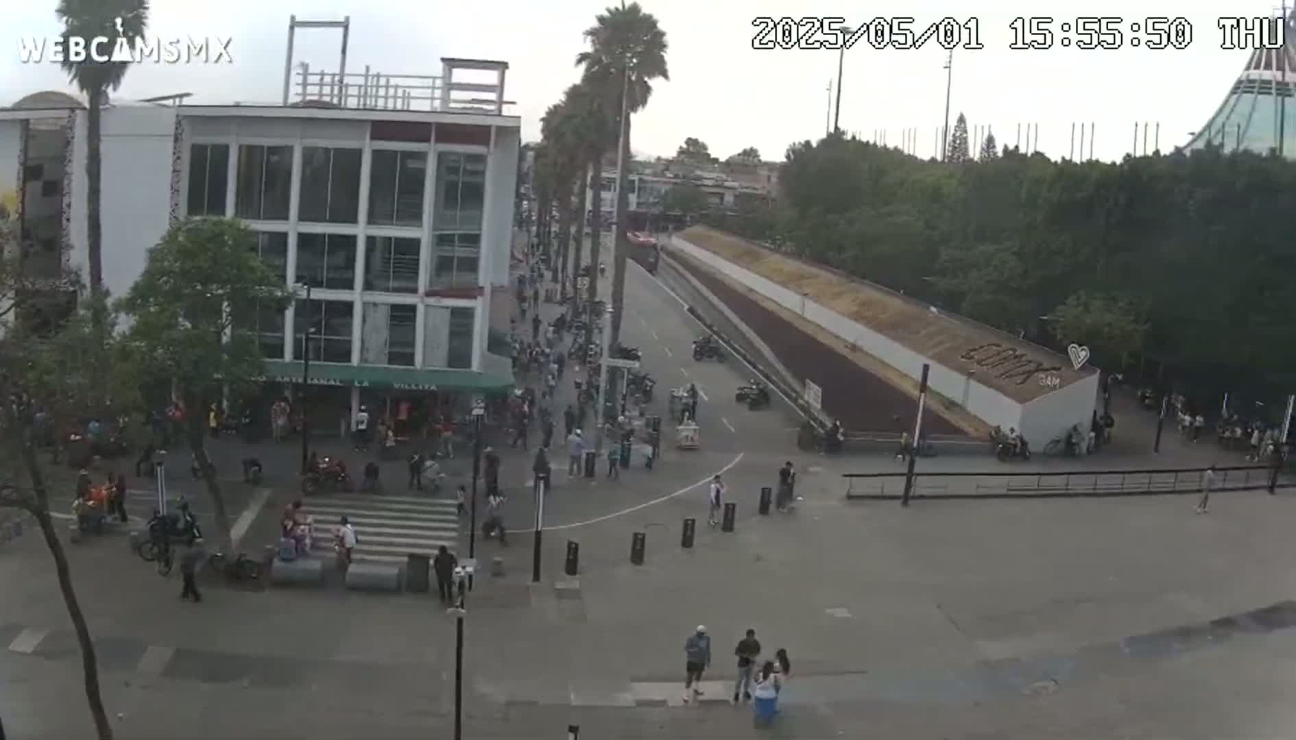 A street scene shows a paved area with pedestrians and parked motorcycles in front of buildings under an overcast sky.