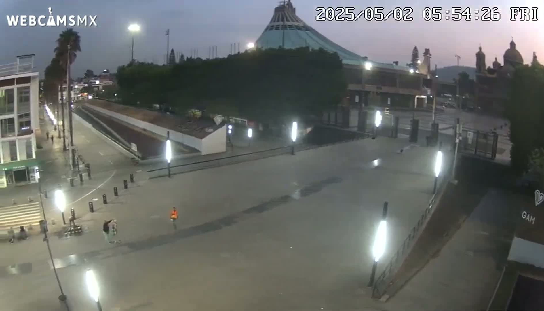 A nighttime, outdoor view of a plaza and large building, illuminated by artificial lights, with a few people visible.