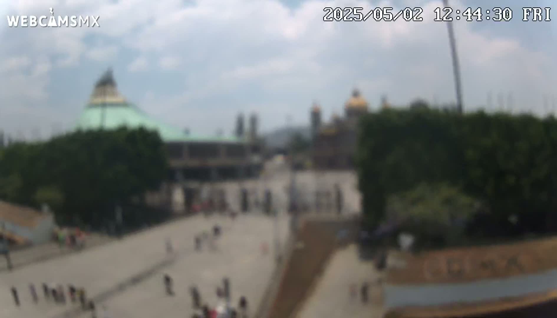 A blurry, wide outdoor shot on a partly cloudy day shows a plaza with several buildings, including a domed structure and a basilica, with many people walking around.
