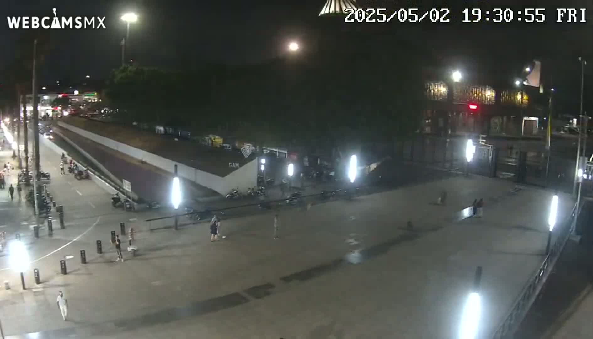 A nighttime, outdoor view of a plaza with a few pedestrians, some parked motorcycles, and bright artificial lighting.