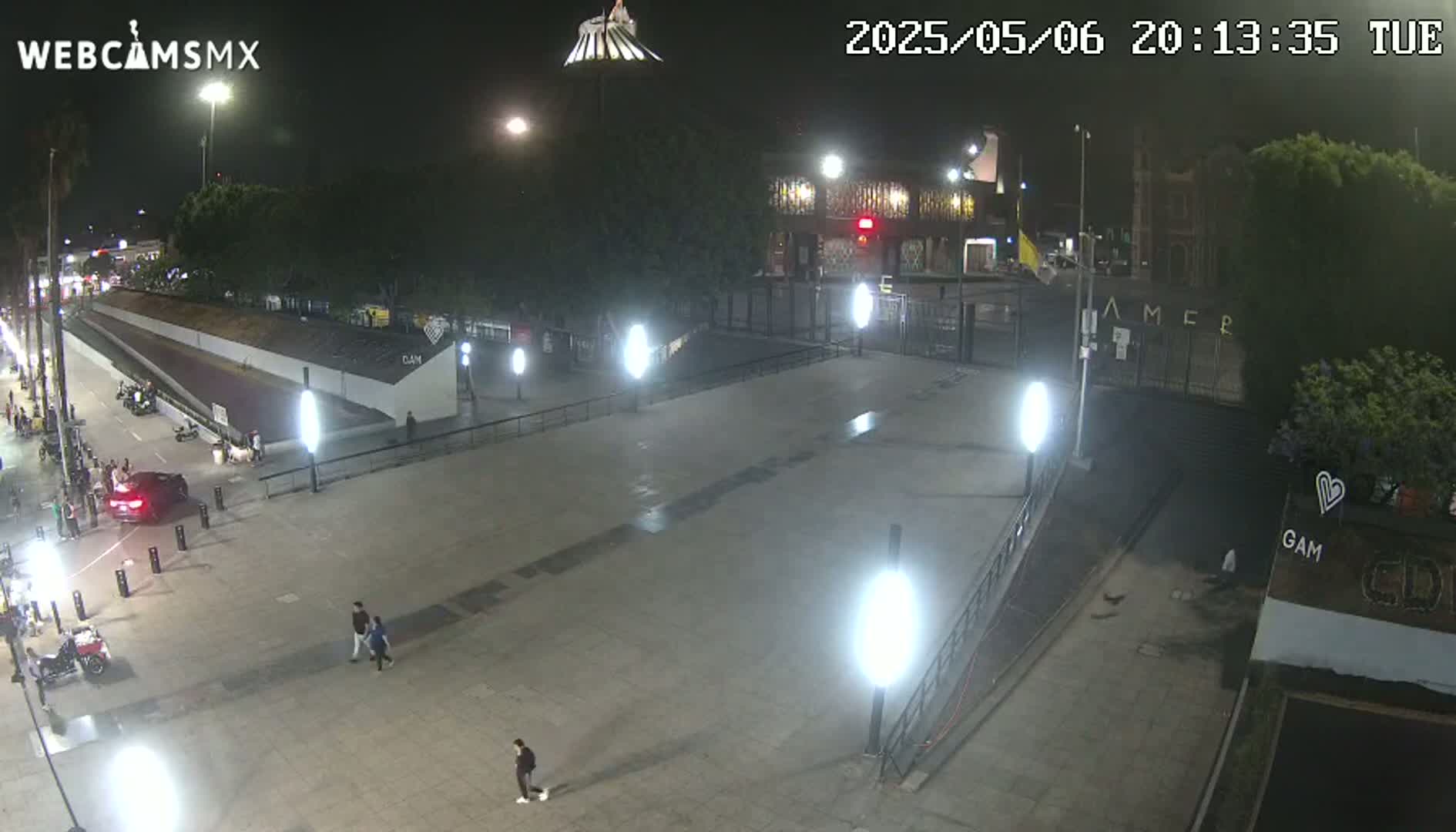 A nighttime, mostly empty plaza with a few pedestrians and cars, is illuminated by streetlights.