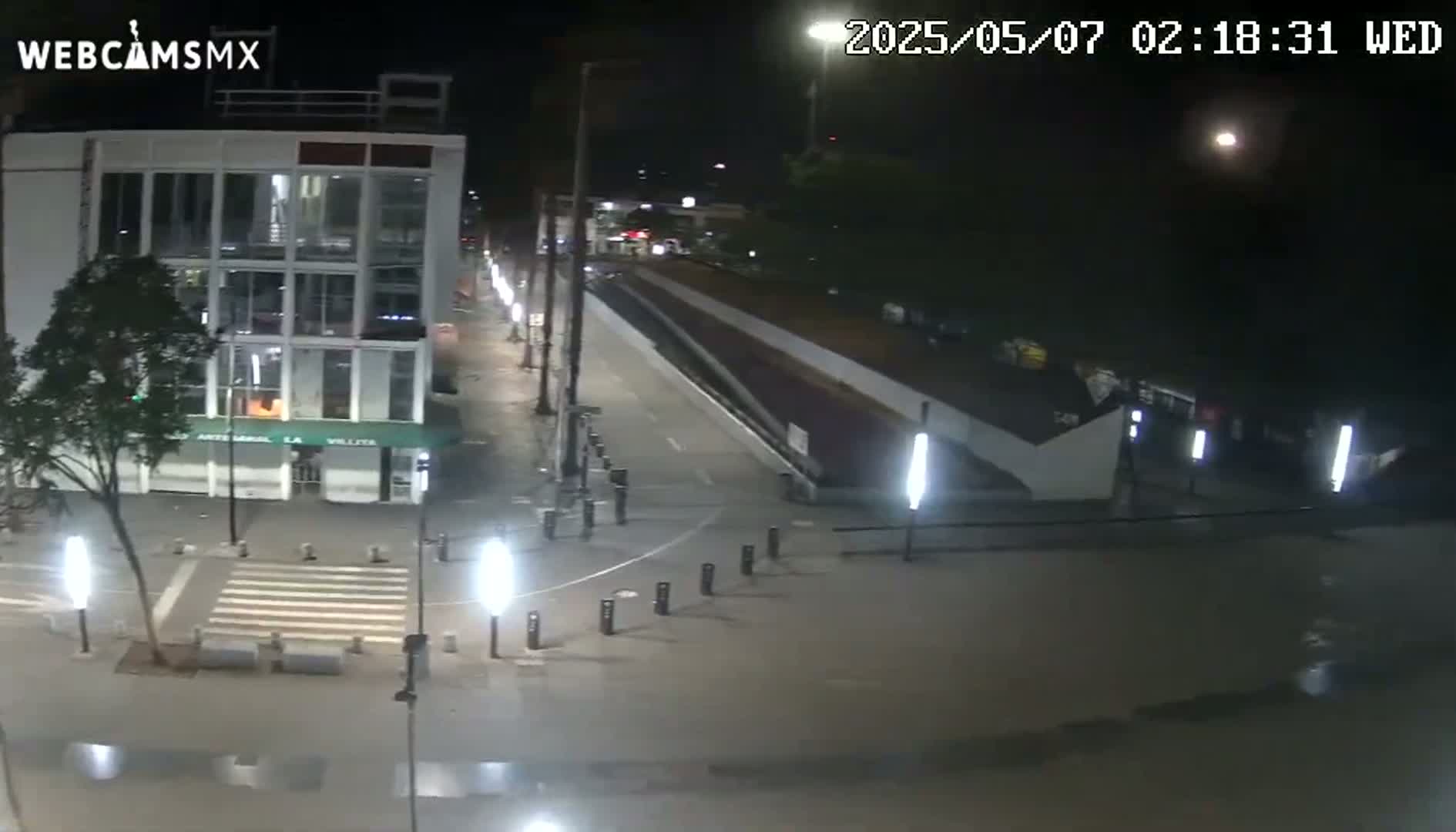 A nighttime, brightly lit street scene shows a multi-story building, a pedestrian crossing, and a mostly empty roadway.