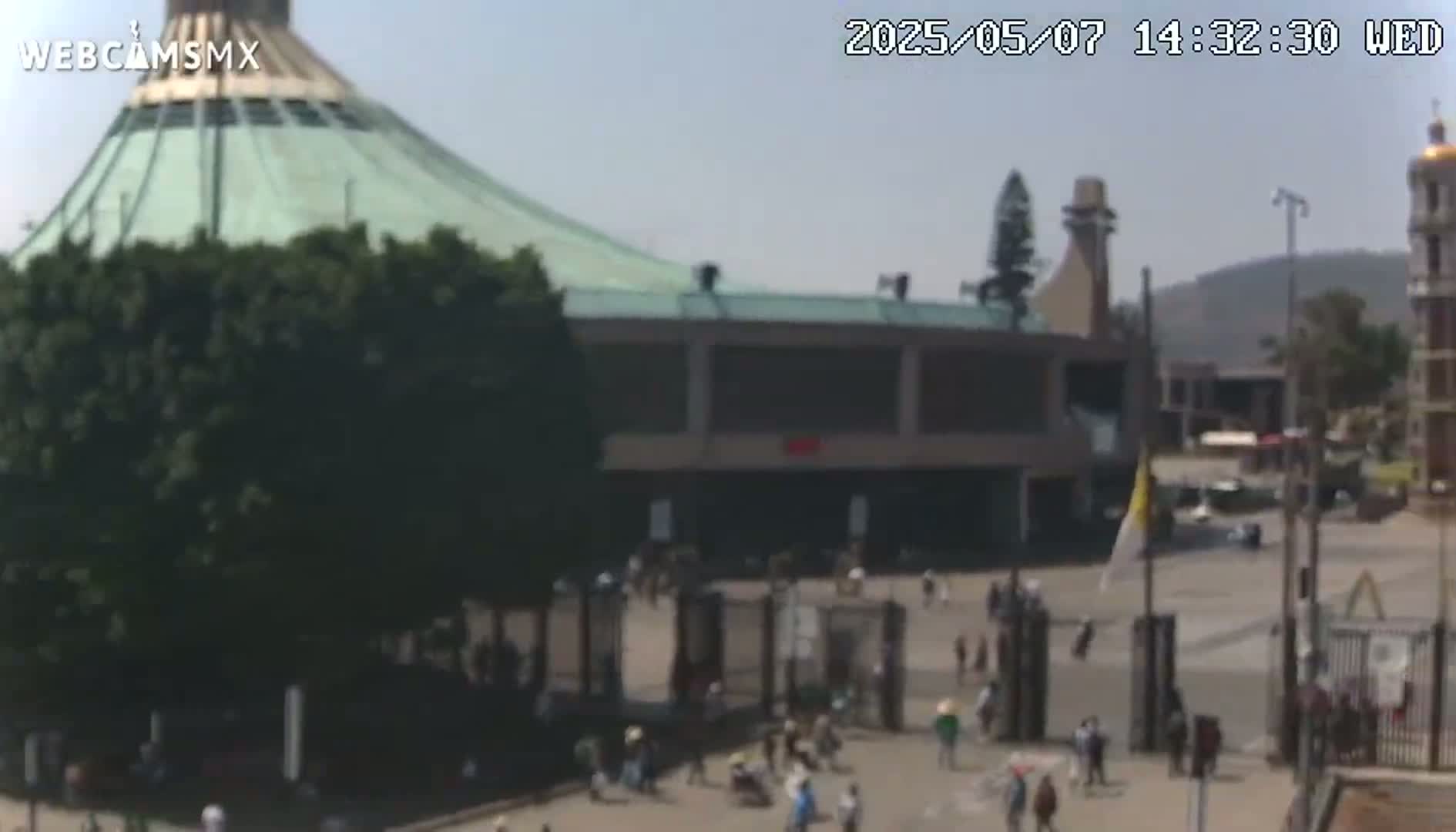 A large, light-green domed building with a plaza full of people in front of it on a clear, sunny day.