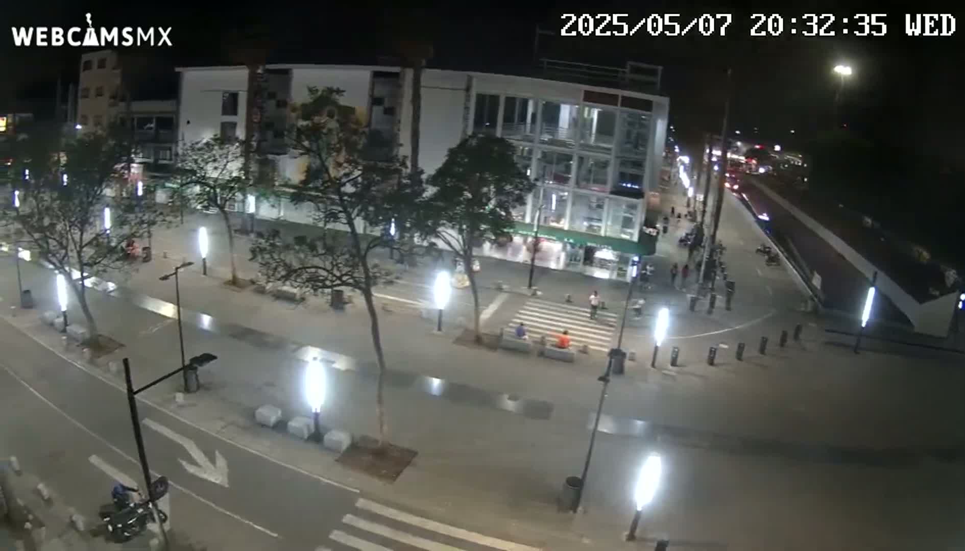 A nighttime, illuminated view of a city street with a few pedestrians and vehicles, showing a modern building and trees lining a paved area.