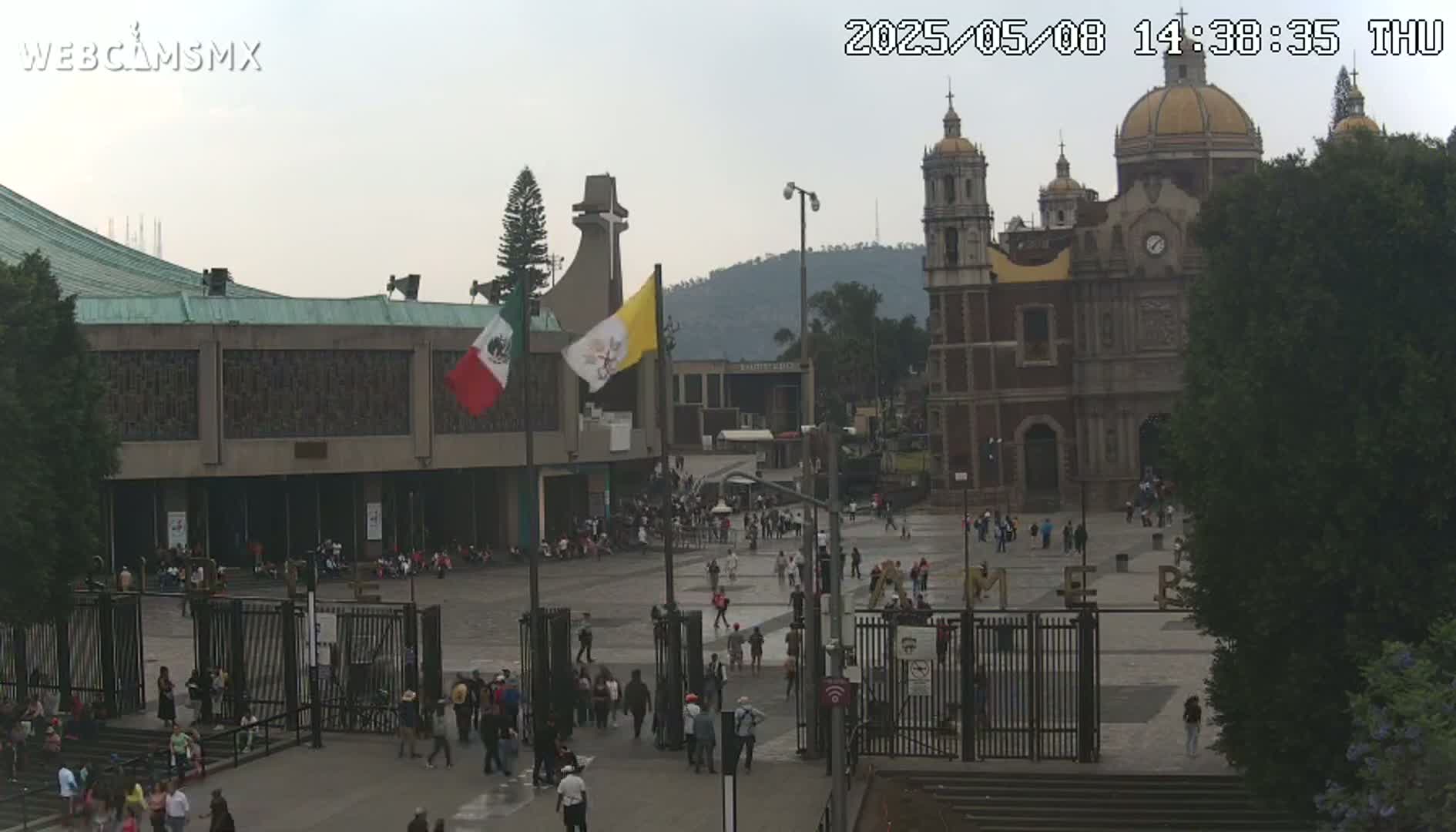 A large plaza with a basilica and other buildings, under an overcast sky, is filled with people.