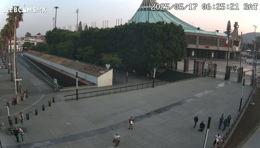 A large plaza with a few pedestrians is in front of a basilica under a mostly clear sky at dawn.