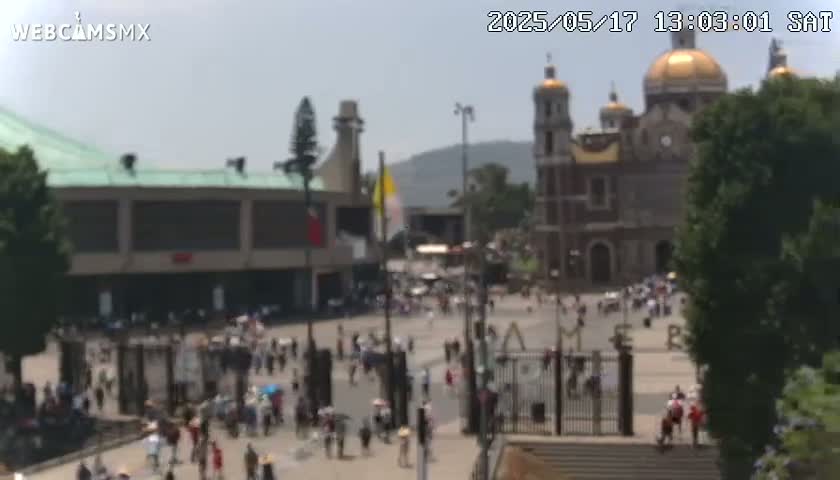A large plaza with many people, a large basilica with golden domes, and other buildings is visible on a hazy, sunny day.