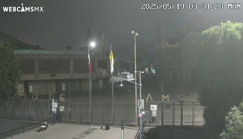 A foggy night scene shows a gated building with flags, streetlights, and a few people near a motorcycle.