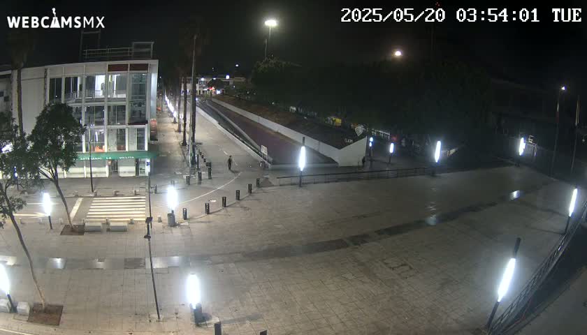 A nighttime view of a city plaza with several buildings, streetlights, and a few pedestrians, under clear skies.
