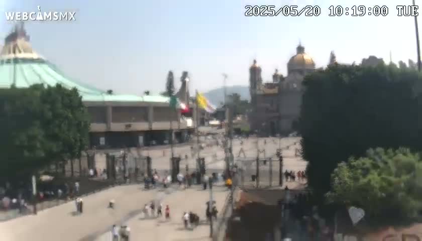 A large plaza with many people, two large buildings, one with a green dome and the other with a gold dome, under a clear, sunny sky.