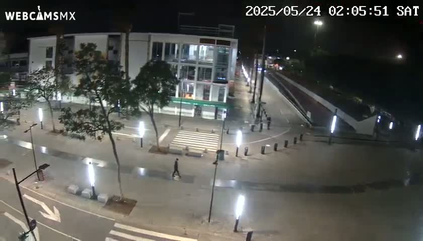 A nighttime, high-angle view of a mostly empty plaza with a multi-story building and a pedestrian crossing, illuminated by streetlights.
