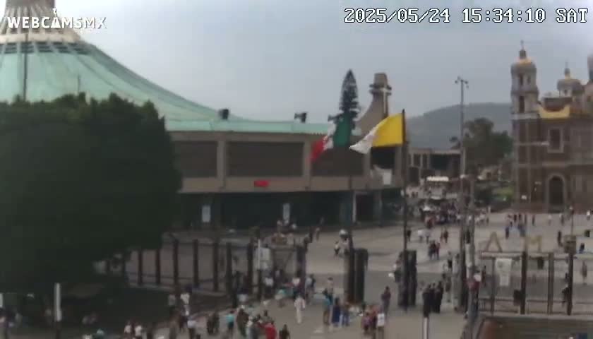 A large plaza with numerous people, surrounded by buildings including a basilica and a smaller church, under an overcast sky.