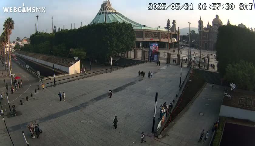 A large plaza with a modern basilica and a smaller, older church in the background, under a mostly clear sky.