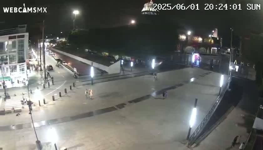 A nighttime, mostly empty plaza with several people scattered about, illuminated by streetlights and building lights.
